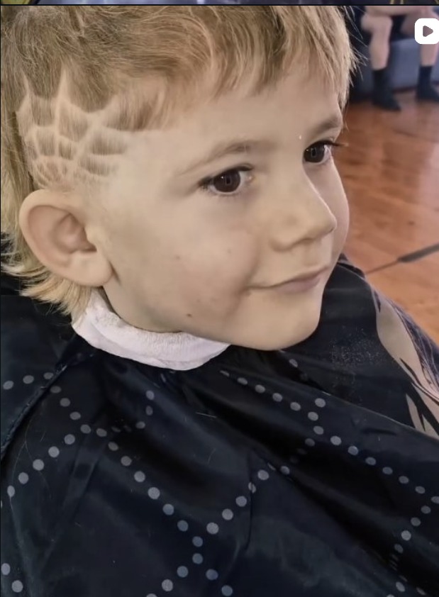 Young boy with a fresh mullet and spiderweb design, smiling in the chair